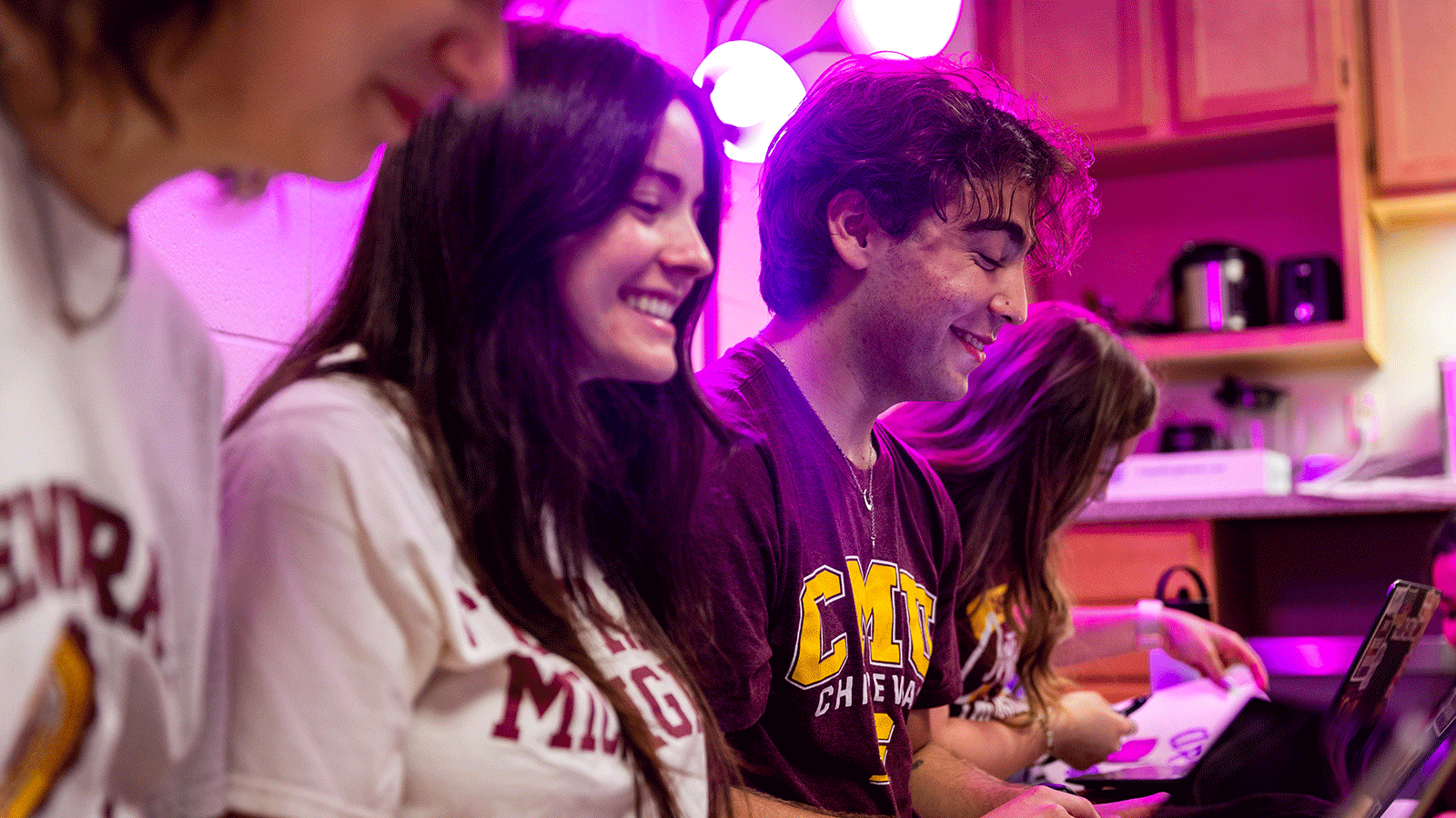 Image of five students sitting on a couch in a residence hall room as they wear CMU t shirts and are working on their laptops, smiling. There is a pink light behind them.