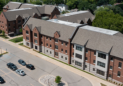 Aerial view of Graduate Housing apartments on the campus of Central Michigan University, captured from a drone. The residence hall is surrounded by lush green grass and trees, with clear blue skies above.