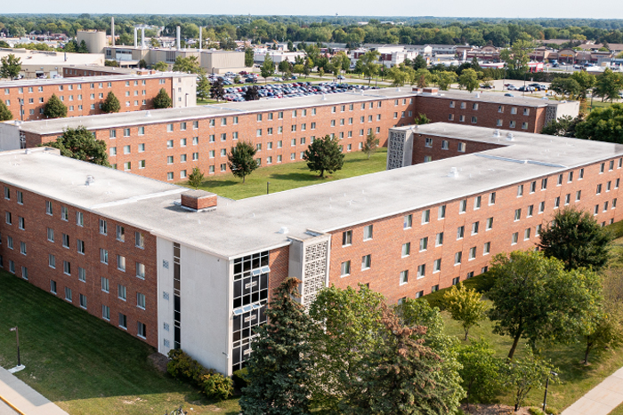 Aerial view of Herrig Hall on the campus of Central Michigan University, captured from a drone. The residence hall is surrounded by lush green grass and trees, with clear blue skies above.