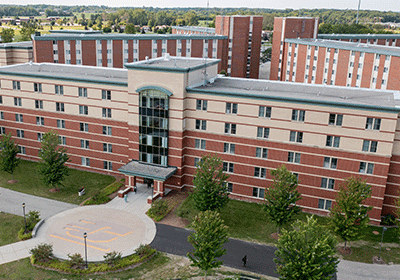 Aerial view of Kesseler Hall on the campus of Central Michigan University, captured from a drone. The residence hall is surrounded by lush green grass and trees, with clear blue skies above.