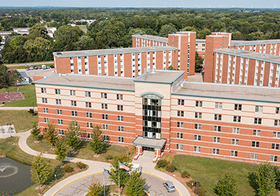 Aerial view of Kulhavi Hall on the campus of Central Michigan University, captured from a drone. The residence hall is surrounded by lush green grass and trees, with clear blue skies above.