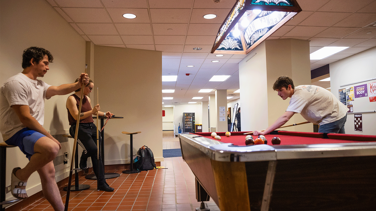 Three students standing around a pool table and playing pool while chatting in a residence hall at Central Michigan University.