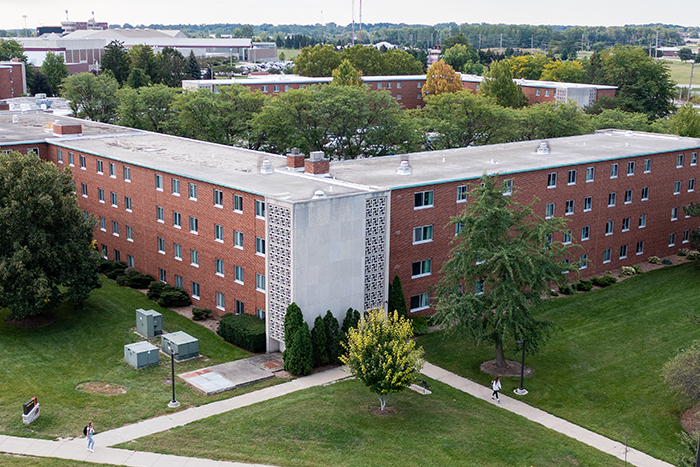 Aerial view of Merrill Hall on the campus of Central Michigan University, captured from a drone. The residence hall is surrounded by lush green grass and trees, with clear blue skies above.