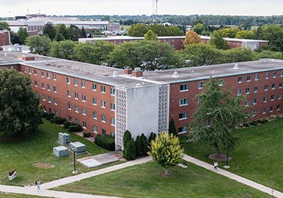 Aerial view of Merrill Hall on the campus of Central Michigan University, captured from a drone. The residence hall is surrounded by lush green grass and trees, with clear blue skies above.