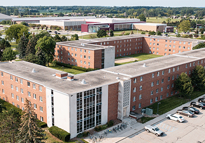 Aerial view of Saxe Hall on the campus of Central Michigan University, captured from a drone. The residence hall is surrounded by lush green grass and trees, with clear blue skies above.