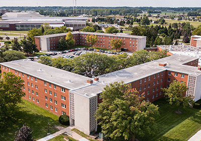 Aerial view of Sweeney Hall on the campus of Central Michigan University, captured from a drone. The residence hall is surrounded by lush green grass and trees, with clear blue skies above.