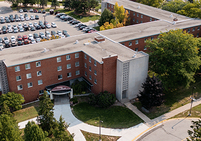 Aerial view of Thorpe Hall on the campus of Central Michigan University, captured from a drone. The residence hall is surrounded by lush green grass and trees, with clear blue skies above.