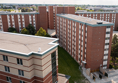 Aerial view of Troutman Hall on the campus of Central Michigan University, captured from a drone. The residence hall is surrounded by lush green grass and trees, with clear blue skies above.