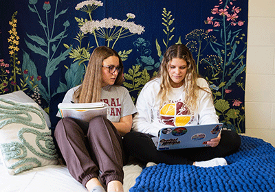 Two students sitting on a bed looking at a laptop in Fabiano Hall.