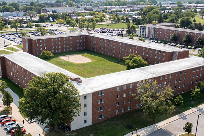 Aerial view of Woldt Hall on the campus of Central Michigan University, captured from a drone. The residence hall is surrounded by lush green grass and trees, with clear blue skies above.