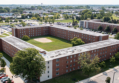 Aerial view of Woldt Hall on the campus of Central Michigan University, captured from a drone. The residence hall is surrounded by lush green grass and trees, with clear blue skies above.