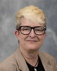 Brie Walter, Director of Sexual Aggression Services profile portrait, wearing black framed glasses, black shirt with beige jacket, and a grey background