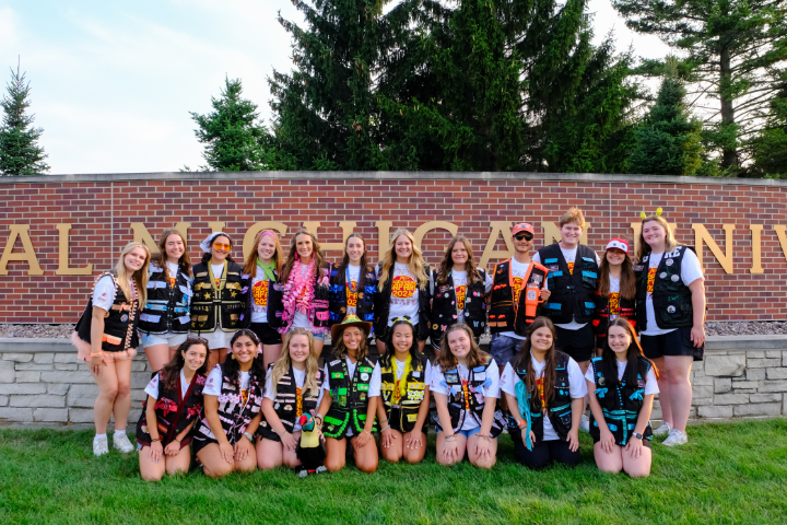 Safari guides are lined up in their vests in front of the Central Michigan University sign smiling and posed