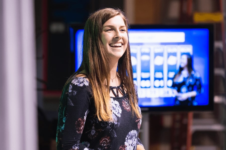 A woman smiling in front of a television delivering news.