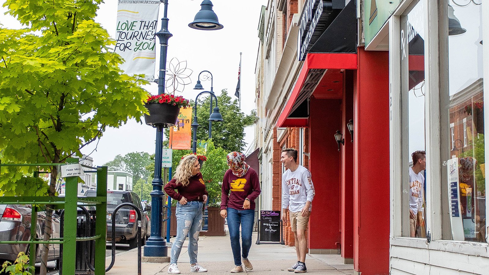 Three students stand on a downtown city sidewalk laughing. The sidewalk has small shops lining the street.