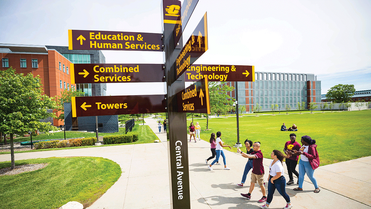 Students walking together through campus with green grass around them as they walk by a sign that has points in various directions that has building names and an action C on it.