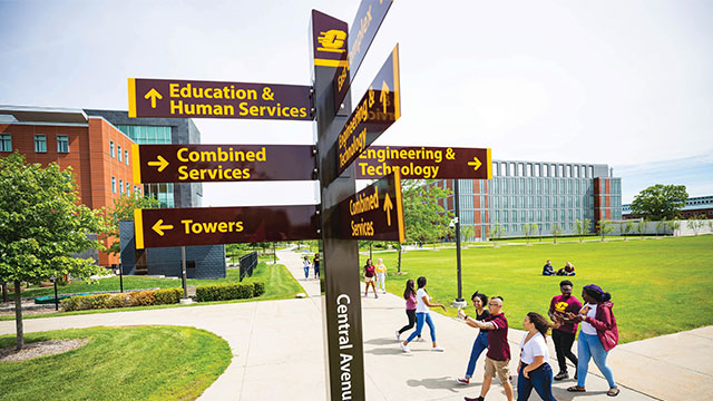 Students walking around campus during orientation. There's a sign pointing to various places on campus such a building and dorms. 