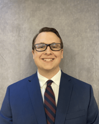 A man in glasses wearing a suit and tie smiles for a headshot.