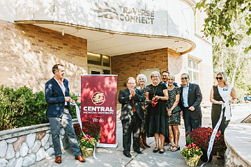 With the Traverse Connect building in the background, 6 CMU officials stand as the ribbon is cut.  Two Traverse Connect officials are holding the ribbon while Betty Kirby is holding the scissors.