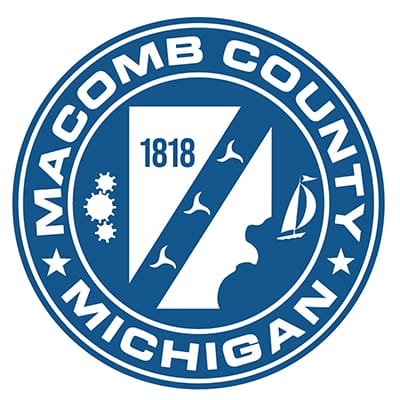 a blue and white circular logo with the words Macomb County Michigan in white in the center is a shape of the county in white and the number 1818 in blue.  On each side of the county are three gears and a sailboat.