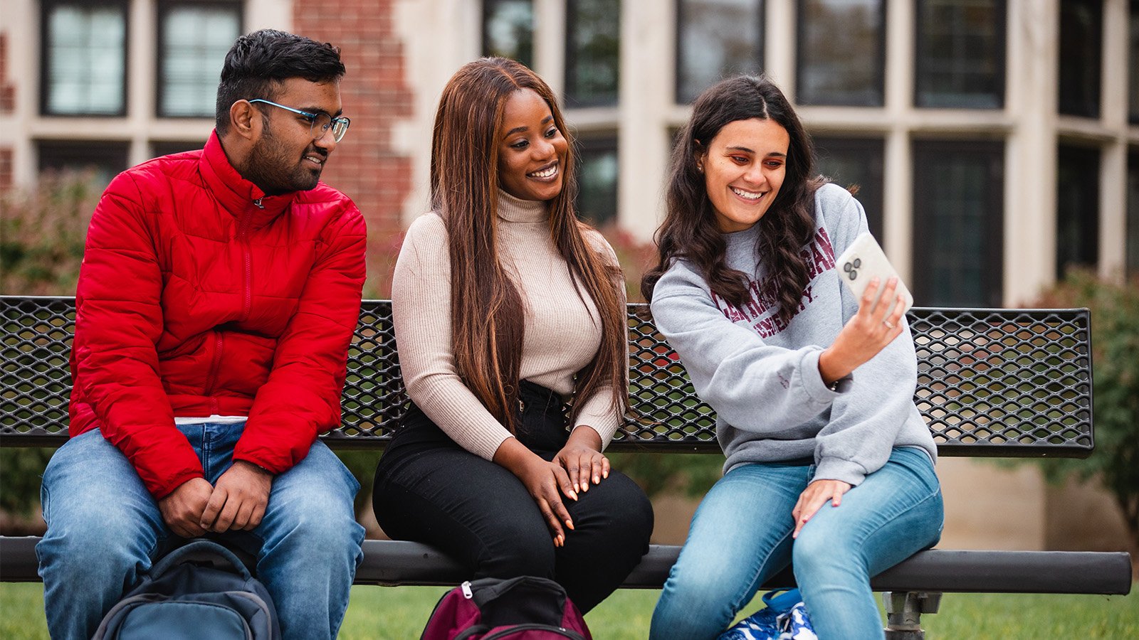 Three students sit on a campus bench together, smiling and posing for a selfie-style photo