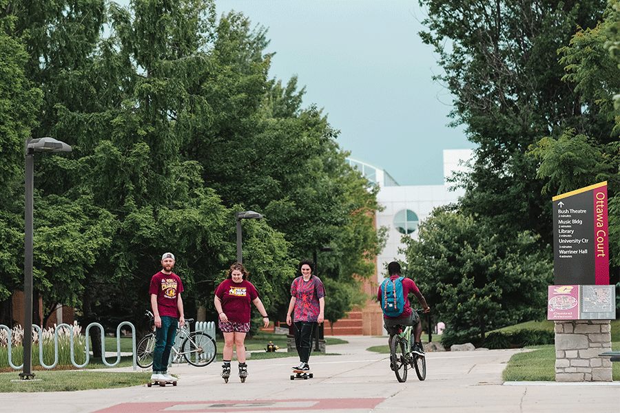 Four students in the middle of campus, one riding a bike, two skateboarding and another on rollerblades. All of them are wearing maroon t-shirts.