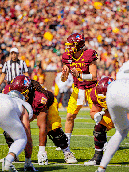 A CMU football player ready to receive the football, wearing maroon and gold, helmet and shoulder pads on a green field surrounded by fans in the distance.