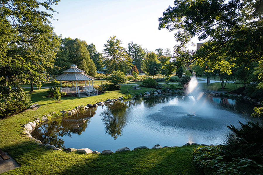 A white gazebo with flowers and grass in the foreground, trees and the sun rising in the background on campus.