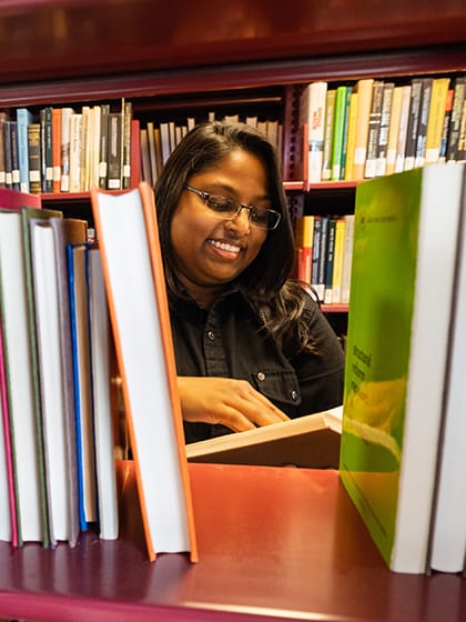 A student with dark hair and glasses looking at a book in the library on campus.