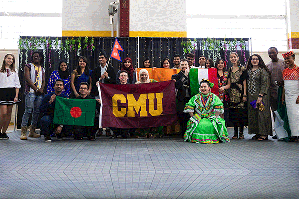A group of students pose around a maroon and gold CMU flag as they are dressed to perform in the international expo.