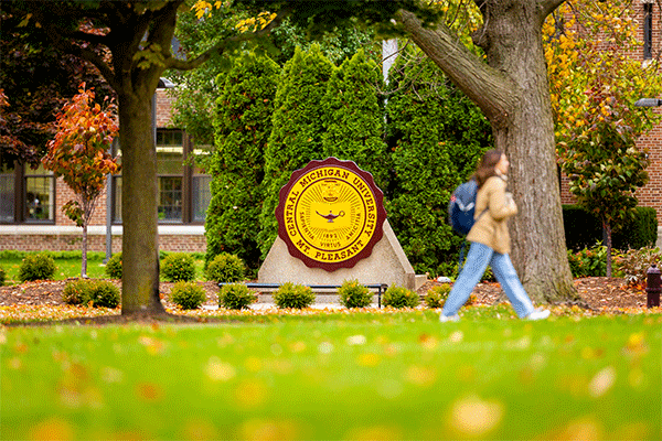 A student walks by the maroon and gold CMU seal statue outside, on campus. There are green shrubs behind the seal, as well as two trees and orange leaves on the green grass.