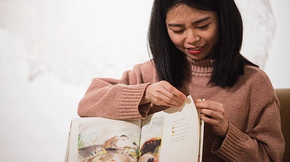 A woman sitting and holding a children's book as she reads it to an audience at the annual Clarke Historical Library, "International Children's Books" event.