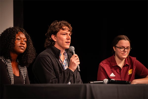 Two students sitting on stage next to a staff member in a maroon polo shirt. The male student is holding a microphone and speaking.