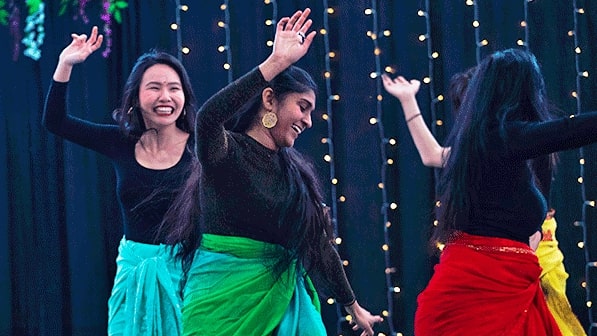 Three students seen dancing on stage at the International Student Expo while wearing black long-sleeved tops, and bright skirts that are blue and green. red and yellow.