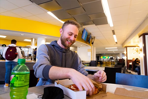 A young man with blonde hair sits at a table holding a small pizza from campus dining options.