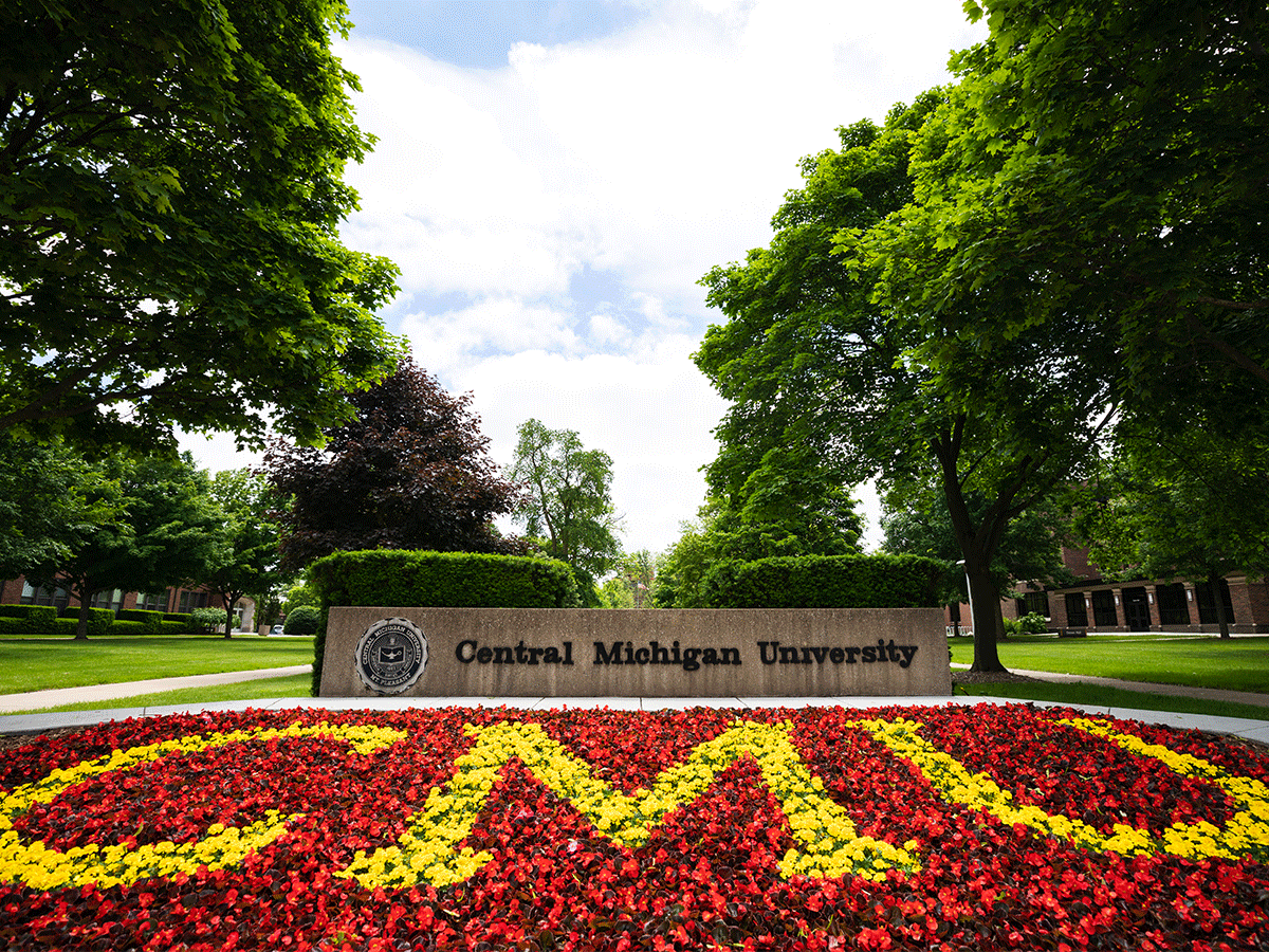 Close up of the Central Michigan University stone sign, with a bed of red and yellow flowers that spell CMU, blue sky and green trees are in the background on a spring day in Michigan.