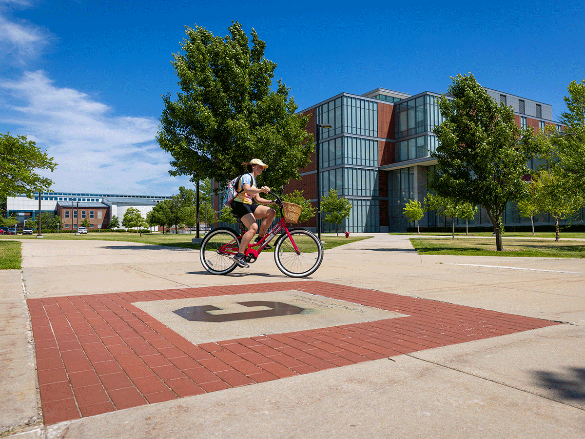 A warm sunny day in the summer in Michigan, a student rides a red bike through campus and passes the Action C plaque in cement. The sky is blue and the trees around are green.