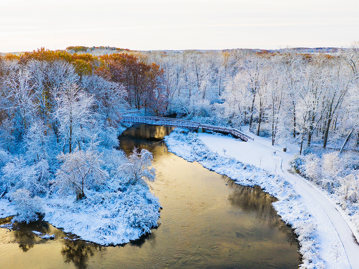 A birds eye view of Mount Pleasant, Michigan's park near campus, showcasing the Chippewa River and a bridge on a cold day in the Winter-all of the trees and ground are covered in white snow.
