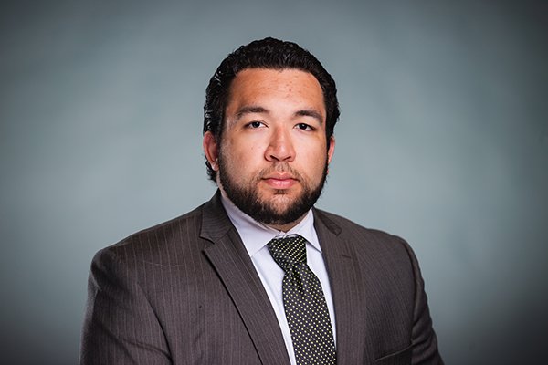 Professional headshot of Max Logan wearing a grey pinstriped jacket, white shirt, and black tie with small yellow polka dots against a light grey background.