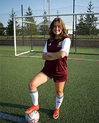 SI Leader for HSC 308 Brooklyn Purdom posing for a photo while standing with one foot on a soccer ball, in a soccer uniform, in front of a soccer goal post.