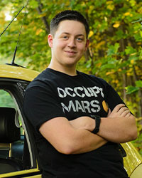 SI Leader Daniel Thanasiu smiling and posing for a photo while standing outside against a yellow car with green trees in the background.