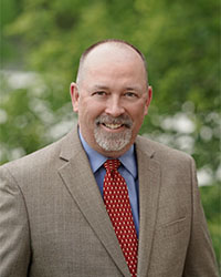 Headshot of Alumni Association Board of Directors member Chris Cantrell standing outside wearing a tan suit with a red spotted tie and a light blue collared shirt and he has a beard and is smiling with teeth.