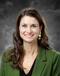 Headshot of Alumni Association Board of Directors member Kelly Eldracher who has long brown curled hair and is smiling with teeth while wearing an olive green blazer and black top.