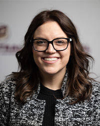Headshot of Alumni Board of Directors Vice President Erica Romac wearing a white and black blazer over a black top and she has black glasses and shoulder-length brown hair is smiling with teeth in front of a CMU backdrop.