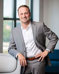 Headshot of Alumni Association Board of Directors member Griffith Gatewood who has blue eyes and short light brown hair wearing a light gray suit and white collared shirt leaning on a gray chair as he smiles with teeth.