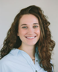 Headshot of Alumni Association Board of Directors member Jennifer Peacock wearing a light blue button down shirt and she has long curly brown hair and is smiling with teeth.