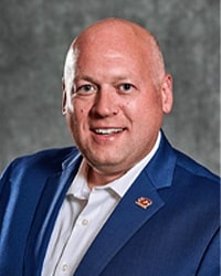 Nate Tallman is an Alumni Board member who holds Emeritus status and was the former president of the Board and he is wearing a blue suit with an Action "C" pin and  a white button-down shirt.