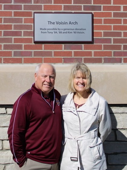 Tony and Kim Voisin standing in front of a sign for The Voisin Arch.