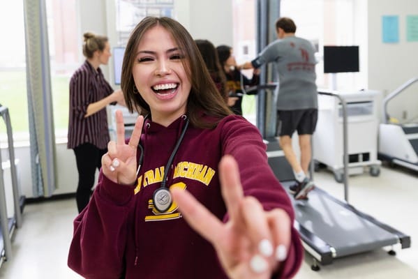 CMU student smiling and holding up two fingers in front of person on treadmill.