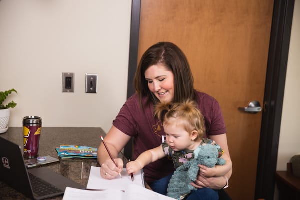 A woman sits at a desk, holding a baby while studying.